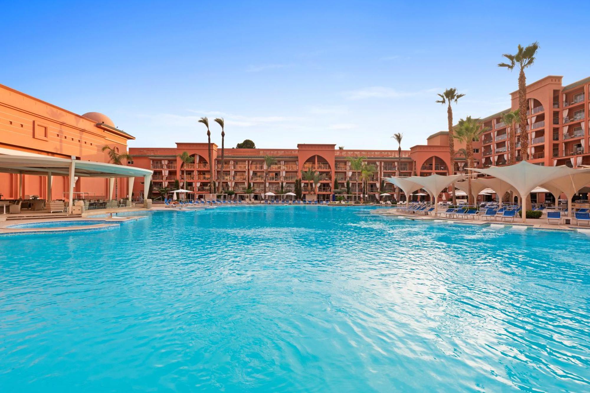 サヴォイ ル グランド ホテル マラケシュ エクステリア 写真 The photo shows a large swimming pool surrounded by a resort. The pool is clear and blue, with lounge chairs and shaded areas nearby. In the background, there are several buildings of the resort, featuring architectural elements like arches and balco